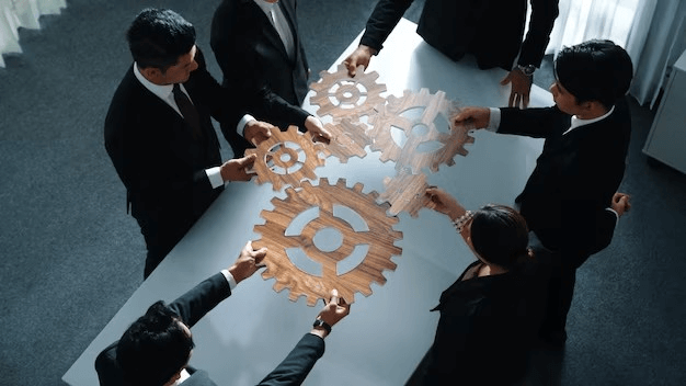 Aerial view of six professionals in suits collaborating, holding wooden gear pieces above a conference table, symbolizing teamwork.