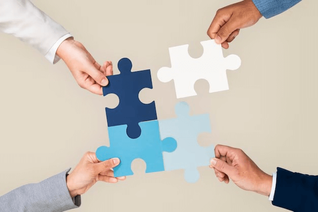 Four hands grasp different puzzle pieces in blue and white, symbolizing teamwork and collaboration against a neutral background.